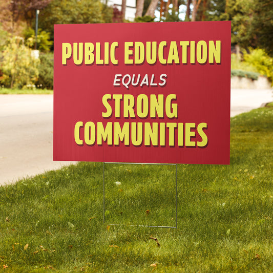 Red sign with yellow text 'PUBLIC EDUCATION EQUALS STRONG COMMUNITIES' on a grassy lawn.